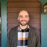 Wayland McQueen wearing an orange, white and black plaid button-up shirt and brown cardigan over an emerald tshirt in front of a brown and green wall