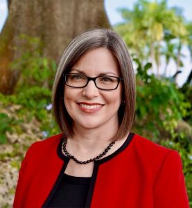 Woman wearing a red jacket and glasses smiling in front of greenery