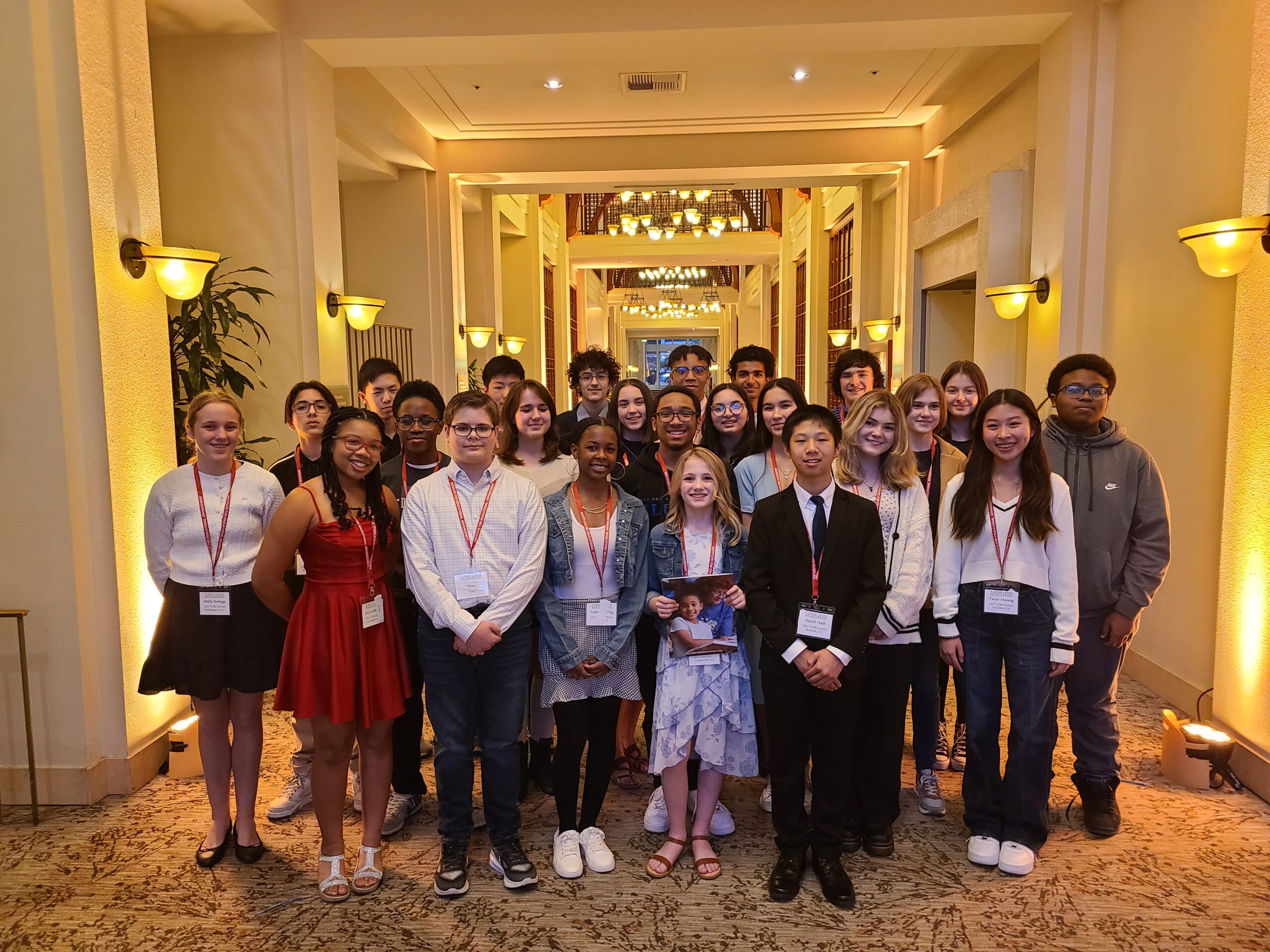 group of 27 8th grade students in semi-casual attire with red lanyards around their necks, smiling together in a large, high-ceiling hallway of a hotel