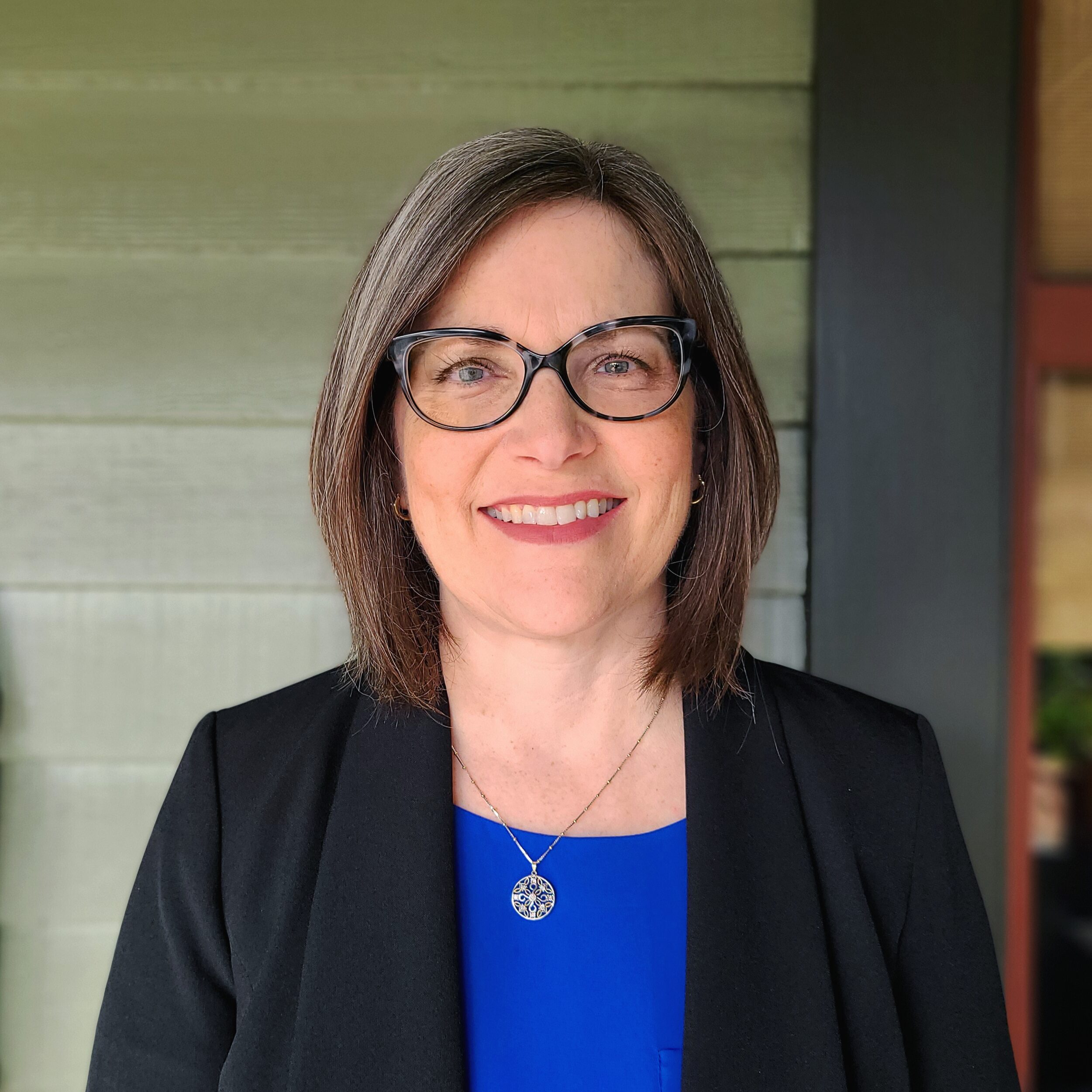 woman with a bob and glasses in a blue shirt and black blazer jacket in front of a green paneled house