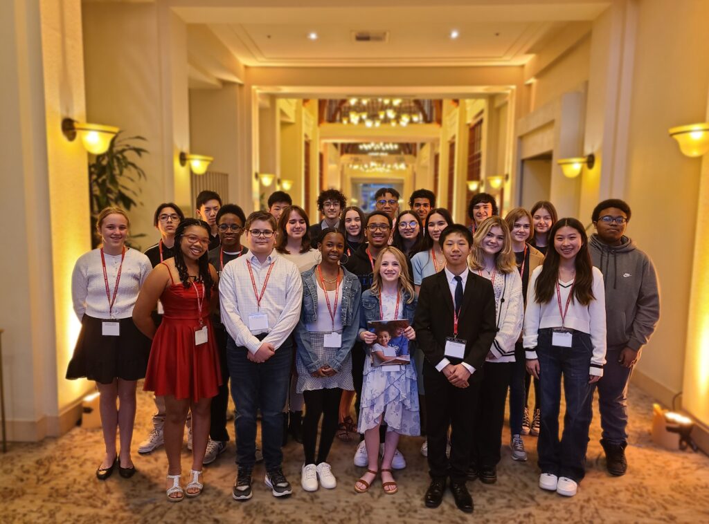 about 30 high school freshmen in dressy-casual attire smiling together, standing in 3 rows in the middle of a hotel lobby with high ceilings