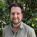 Andy in an olive button down shirt in front of large green hedge